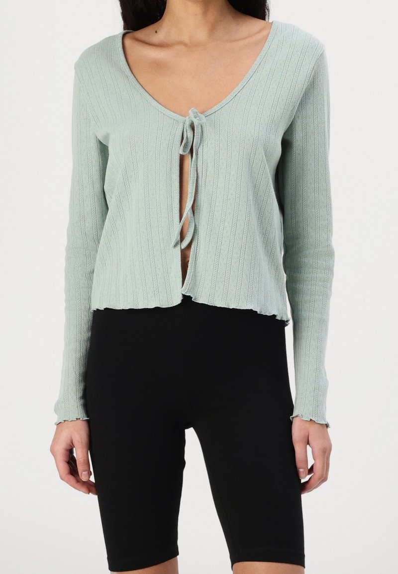 Cardigan vert clair côtelé avec un décolleté en V profond et une fermeture à nouer, avec un ourlet effiloché et des manches longues, associé à un short noir.