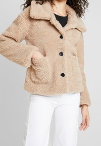 Osoba má na sobě béžovou, huňatou bundu s límcem a kapsami, stylizovanou s bílými kalhotami, stojící na jednobarevném pozadí.