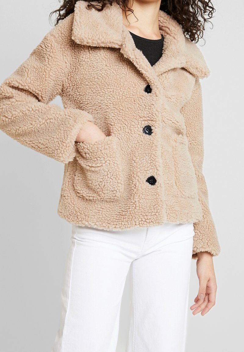 Osoba má na sobě béžovou, huňatou bundu s límcem a kapsami, stylizovanou s bílými kalhotami, stojící na jednobarevném pozadí.