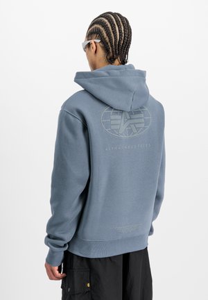 Person mit geflochtenem Haar, der einen hellblauen Hoodie von Alpha Industries und schwarze Cargo-Hosen trägt, steht mit dem Rücken zu einem schlichten Hintergrund.