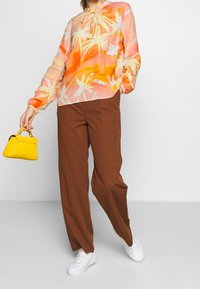Lekka, przezroczysta bluzka z nadrukiem palm w kolorze pomarańczowym i różowym, zestawiona z brązowymi spodniami o szerokich nogawkach i białymi trampkami. Akcent w postaci żółtej torby.