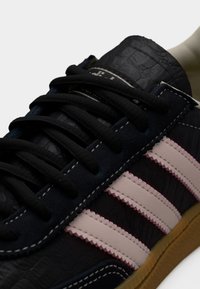 Κοντινό πλάνο μαύρου αθλητικού παπουτσιού με υφή από δέρμα, μαύρα κορδόνια, τρεις μπεζ ρίγες στο πλάι, λευκά ραφές και σόλα από καουτσούκ.