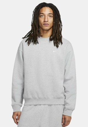 Sudadera gris con cuello redondo, mangas largas y puños de canalé. Presenta un pequeño logotipo blanco de Nike en el pecho. Textura suave.