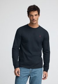 Mörkblå bomullsskjorta med lång ärm och rund halsringning, med en liten röd logotyp på vänster bröst. Tyget verkar slätt och åtsittande.