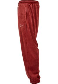 Red velvet joggers with elastic waistband, side pockets, and ribbed cuffs. Features subtle logo accent on the left thigh. Soft texture.