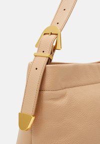 Sac à main en cuir beige avec une finition texturée, arborant une boucle en métal doré et une bandoulière ajustable. Détails de couture visibles près de l'ouverture.