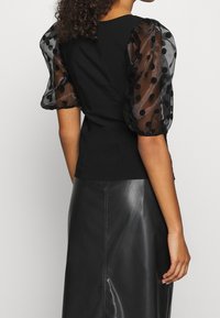 Top nero con maniche trasparenti a sbuffo caratterizzato da pois neri, abbinato a una gonna in pelle nera, design elegante e aderente.