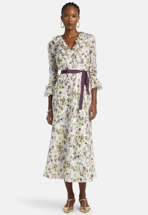 Kvinde iført en hvid midikjole med blomsterprint, flæsekant ved kraven og ærmerne, bundet med et lilla bælte, kombineret med gyldne hæle.