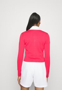 Rosa stickad cardigan med långa ärmar, ribbade mudd och nederkant, kombinerad med vita shorts. Bakifrån visar den en slät yta utan utsmyckningar.