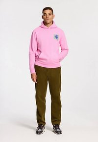 Rózsaszín kapucnis pulóver elülső zsebbel és logó folttal, olíva zöld kordbársony nadrággal és mintás tornacipővel párosítva. Semleges háttér.