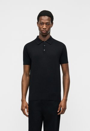 Homme debout devant un fond uni, portant un polo noir ajusté à manches courtes avec trois boutons et un pantalon noir.