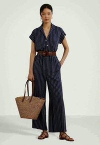 Γυναίκα που φοράει μια ναυτική μπλε οριζόντια ριγέ φόρμα με ένα φαρδύ καφέ ζώνη, κρ�ατώντας μια υφαντή τσάντα και φορώντας καφέ σανδάλια με λουριά.