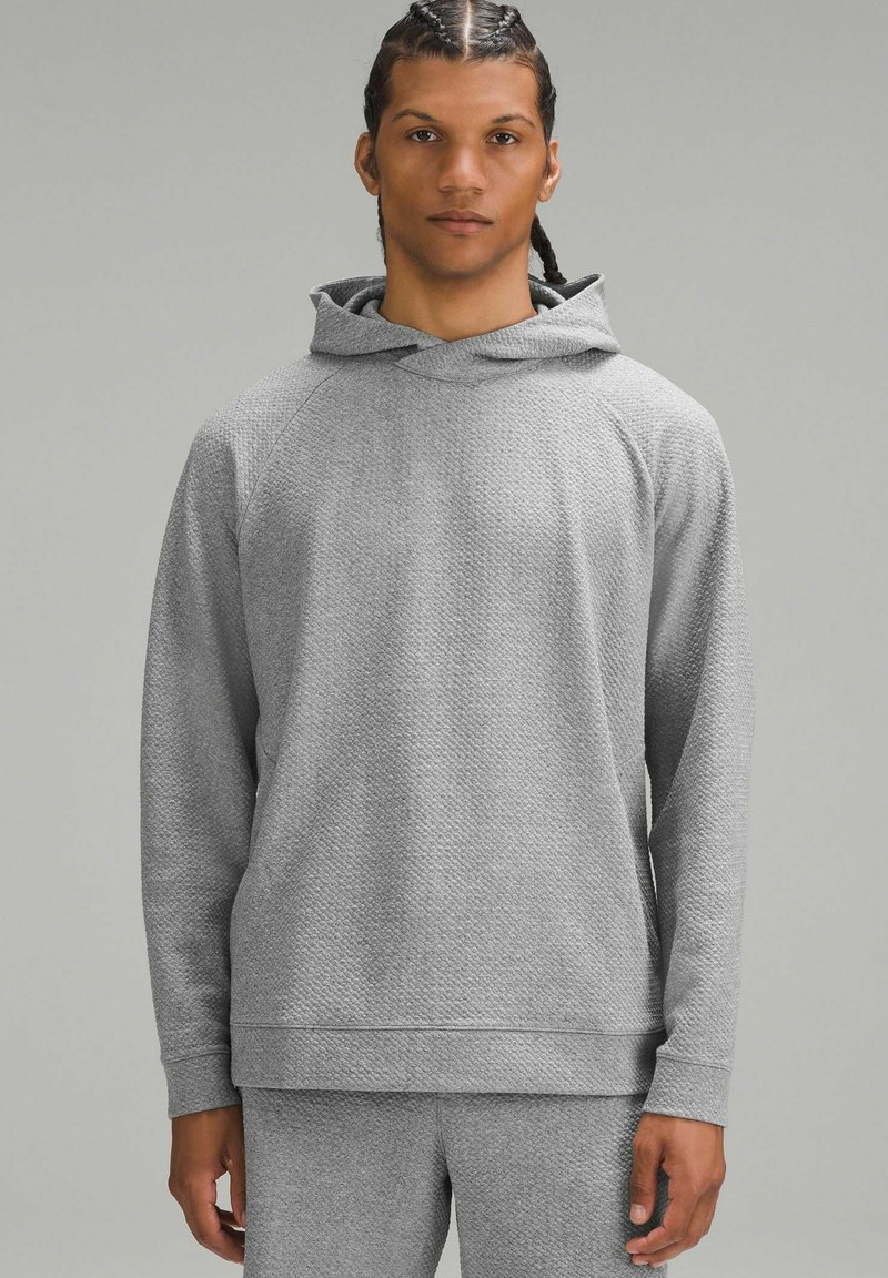 Szary, teksturowany hoodie z przestronnym kapturem, długimi rękawami i luźnym krojem. Tkanina ma subtelny wzór oraz ściągacze przy rękawach.