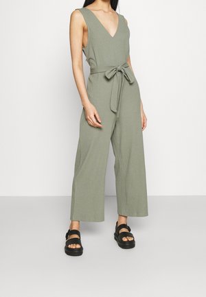 Frau trägt einen ärmellosen, salbeigrünen, gerippten Overall mit weitem Bein und gebundenem Taillengürtel, kombiniert mit schwarzen Plateau-Sandalen.