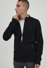 Schwarze Zip-Up-Trainingsjacke mit einem gelben Innenkragen, zwei Vordertaschen mit goldenen Knöpfen und verstellbarem weißen Kordelzug.