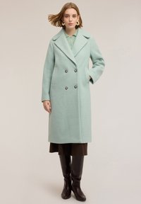 Abrigo verde menta de doble botonadura con tejido texturizado, grandes solapas y botones plateados, combinado con una falda marrón oscuro y botas hasta la rodilla.