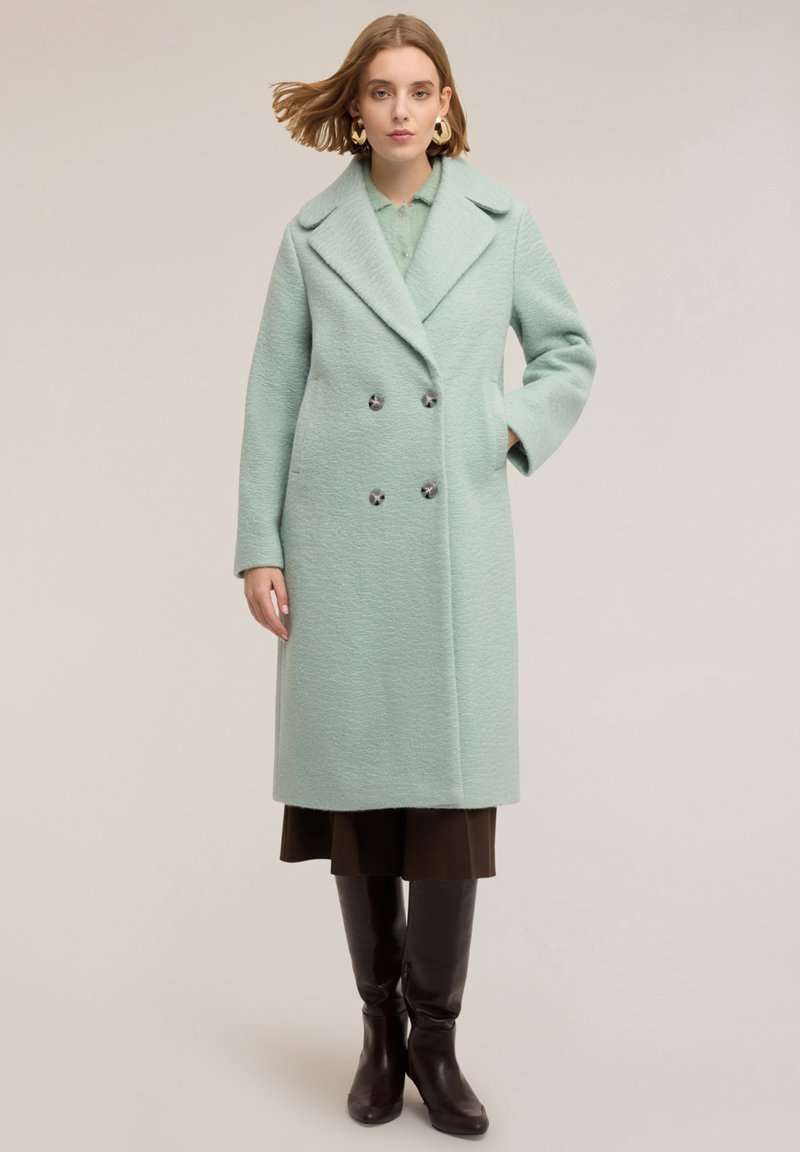 Abrigo verde menta de doble botonadura con tejido texturizado, grandes solapas y botones plateados, combinado con una falda marrón oscuro y botas hasta la rodilla.