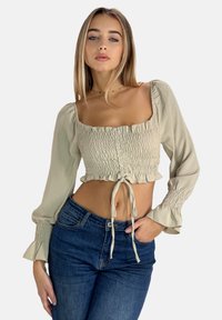 Hellbeige, kurz geschnittene Bluse mit quadratischem Ausschnitt, Schmetterlingsärmeln, gerafftem Material und Bindegürtel in der Taille, kombiniert mit dunkelblauen Jeans.