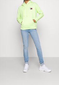 Felpa verde neon con cordini e tasca frontale, abbinata a jeans skinny blu chiaro e sneakers bianche, in piedi di fronte a uno sfondo semplice.