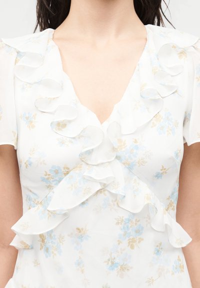 Γυναίκα που φοράει λευκό πουκάμισο με ανοιχτό μπλε και μπεζ floral σχέδιο, σφηκοφωλιά V-λαιμόκοψη με βολάν και λεπτομέρειες μπροστά.