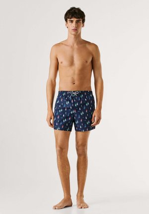 Noor mees paljajalu, kandes tumesiniseid ujumispükse, millel on värvikad purjelaeva mustrid, ereda ühevärvilise tausta ees.