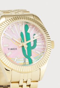 Montre dorée avec une lunette striée, présentant un motif de cactus coloré sur un cadran en nacre rose et des index horaires rectangulaires.