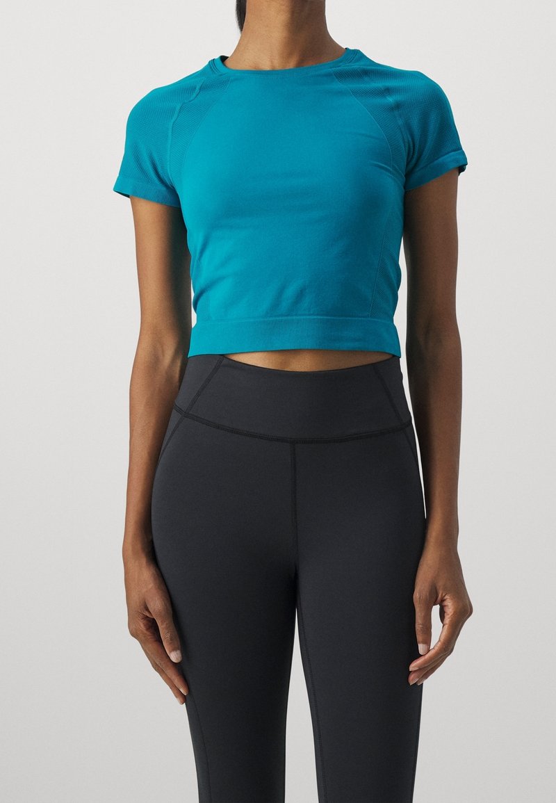 Top atletico corto color teal con maniche corte e texture a coste, abbinato a leggings neri a vita alta con dettagli di cucitura visibili.