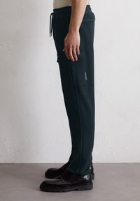 Pantalons de jogging vert foncé en tissu texturé, dotés d'une poche cargo latérale et d'une ceinture élastique. Portés avec des chaussures noires à lacets.