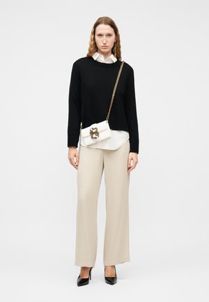 Γυναίκα που φοράει μαύρο πουλόβερ πάνω από λευκό πουκάμισο, μπεζ παντελόνια με φαρδιά πόδια, μαύρες γόβες και λευκή τσάντα χιαστί με χρυσή αλυσίδα και λεπτομέρεια φιδιού.