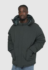 Mørkegrønne dunjakke med hætte, der har et quiltet design, lynlåslommer og elastiske manchetter. Indersiden er foret med et kontrasterende materiale.