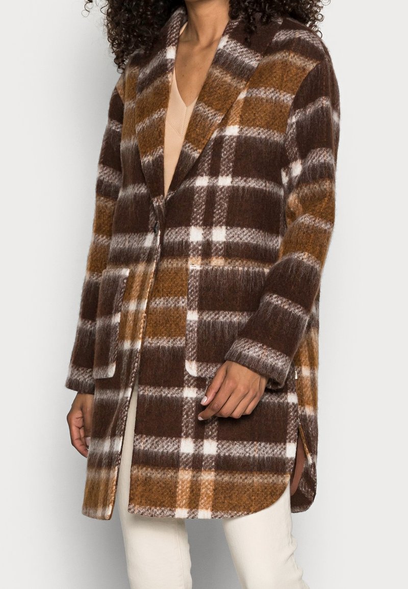 Femme portant un manteau en laine à carreaux marron, orange et blanc avec de grandes poches avant et un col cranté, par-dessus des vêtements beiges.