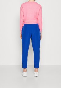 Osoba v jasně růžovém dlouhém rukávu, v úzkých modrých kalhotách a v bílých teniskách, stojící před jednobarevným světlým pozadím.