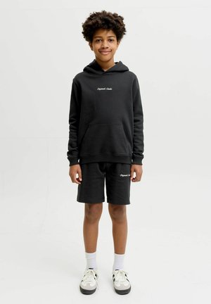 Garçon aux cheveux bouclés portant un sweat à capuche noir et un short avec un texte blanc, des chaussettes blanches et des baskets, debout devant un fond clair.