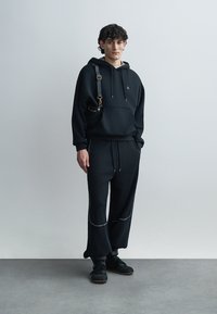 Μαύρο φούτερ με κουκούλα και κορδόνια, και μπροστινή τσέπη, σε συνδυασμό με χαλαρής εφαρμογής παντελόνια σε ταιριαστό χρώμα με ανοιχτό γκρι λεπτομέρειες και πρακτικά παπούτσια.