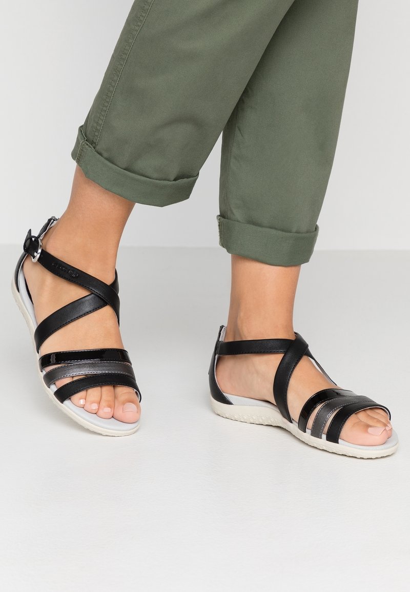 Jalkaterät, joissa on mustat nauhansandaalit valkoisilla pohjilla, yhdistettynä oliivinvihreisiin käärityihin housuihin, seisoo vaalealla pinnalla.