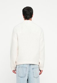 Pulover supradimensionat din tricot crem, cu guler înalt și mansete texturate. Purtați-l peste blugi din denim albastru deschis, evidențiind un material moale și texturat.