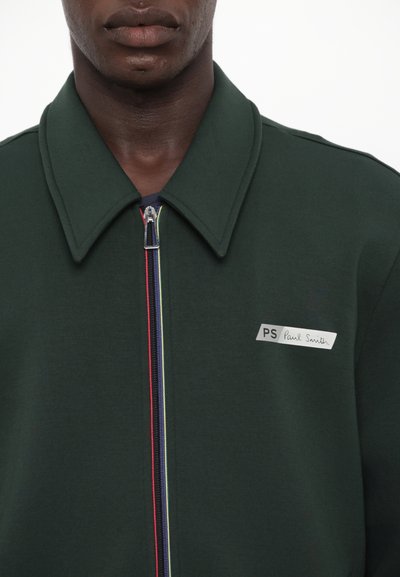 Mørkegrønn zip-up jakke med stor krage, med en flerfarget glidelås og et merket rektangulært patch på venstre side.