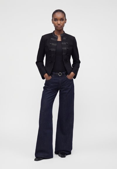 Femme debout les mains dans les poches, portant une veste noire style militaire, un haut noir, un jean large foncé et des chaussures noires sur fond uni.