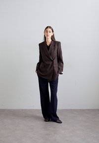 Young woman stands against plain wall wearing oversized dark brown blazer, black top, wide-leg navy trousers, and black mesh shoes.