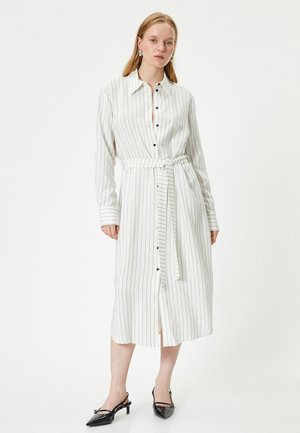Witte gestreepte lange jurk met knoopsluiting, lange mouwen, een ceintuur in de taille en een strikceintuur. Zwarte knopen en puntige zwarte schoenen maken de outfit compleet.