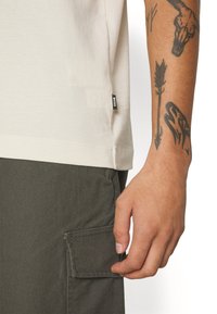 T-shirt en coton beige clair avec un ourlet arrondi, arborant une petite étiquette de logo noire et une poche de pantalon cargo texturée en vert olive.