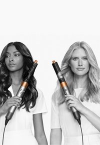 Due donne tengono strumenti per acconciature con un design grigio e argento, caratterizzati da accenti arancioni e barilotti testurizzati. Cavo nero visibile.