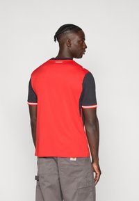 Piros és fekete sportmez rövid ujjakkal. Kontrasztos színű kialakítással, kerek nyakkivágással és kontrasztos csíkozással a mandzsettákon.