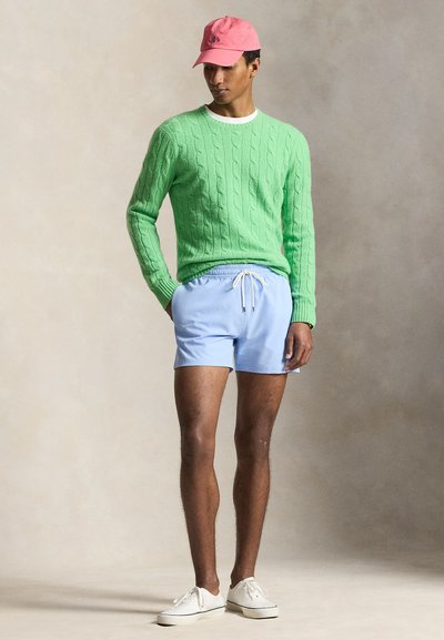 Άνδρας που φοράει πράσινο πουλόβερ πλεχτό με καλώδια, ανοιχτό μπλε σορτς, ροζ καπέλο μπέιζμπολ και λευκά αθλητικά παπούτσια, στέκεται μπροστά σε ουδέτερο φόντο.