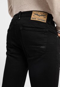 Jean en denim noir avec une coupe droite, une texture subtile et une patch en cuir à la taille avec une marque en relief.