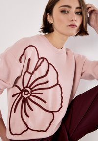 Lichtroze trui met een groot bordeauxrood bloemenontwerp, voorzien van een ronde halslijn en een zachte textuur. De bloem heeft omlijnde details.