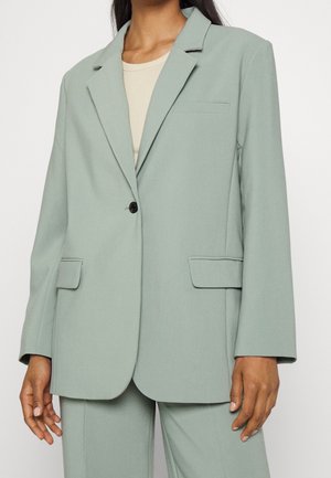 Vrouw draagt een lichtgroene oversized colbert met inkeping revers, één zwarte knoop en klepzakken over een beige top.