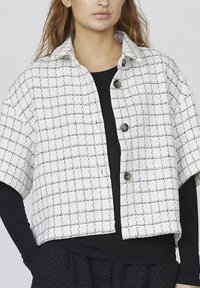 Hvid cropped jakke med sort rutenetmønster, der har en krave og tre mørke knapper. Tekstureret stof med korte ærmer.