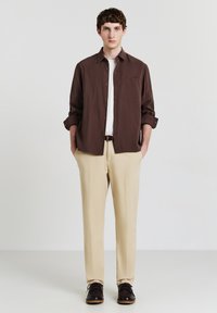 Giovane uomo in piedi con le mani in tasca, indossa una camicia marrone con bottoni, una maglietta bianca sotto, pantaloni beige e scarpe marroni scure su uno sfondo semplice.