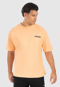 Kortærmet T-shirt i lys ferskenfarvet bomuld med rund hals og sort "SMILODOX" logo på venstre bryst. Enkel design, afslappet pasform.